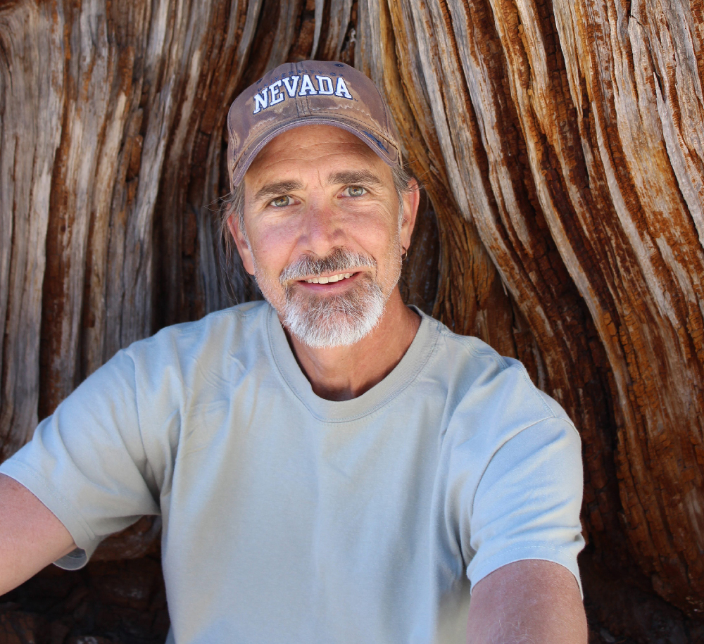 Photo of author Michael Branch, wearing a hat with NEVADA on it, light blue shirt and sitting in front of a the trunk of a very large tree. 