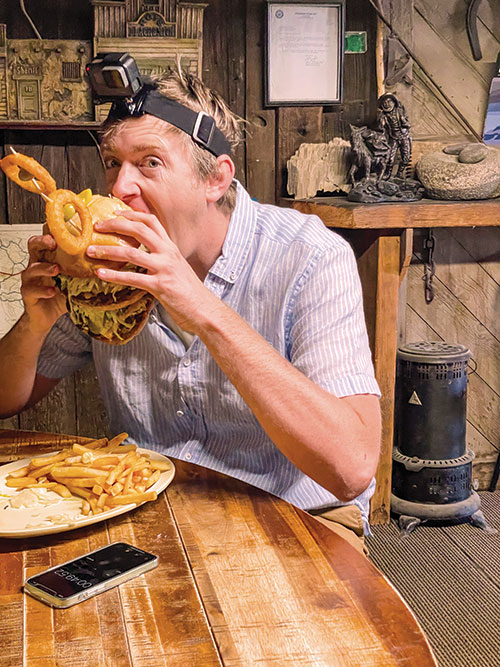 Associate Editor Cory Munson, eating a Monster Burger: Two hamburgers stacked atop one another with a third bun in the middle, with tomato, pickle, and onion. Top bun has a pepperoncini for a nose, and two onion rings with toothpics stuck through them and olives in the middle for eyes. Plate also has a pile of french fries.