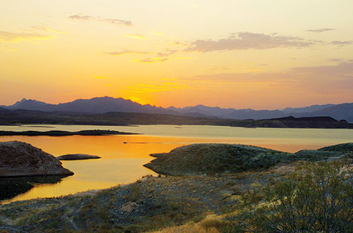 A sunrise or sunset at Lake Mead, with one lone powerboat seen in the water. Mountains in the distance and sagebrush and rabbit brush in the foreground.
