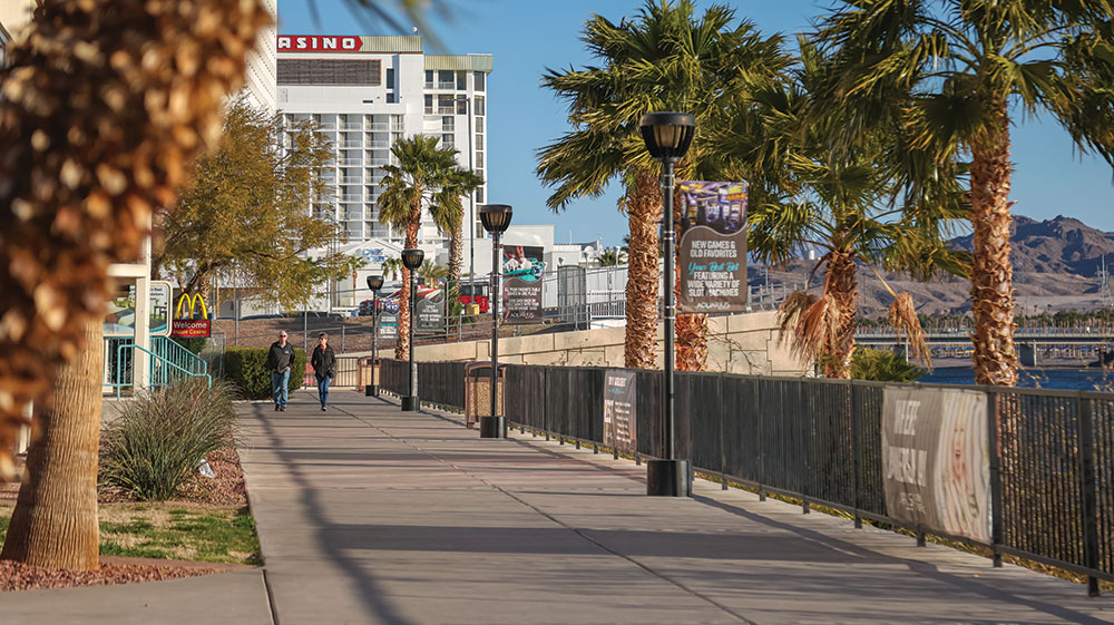 Two people walk on the sidewalk alongside the water, in Laughlin. There are many palm trees, a casino, and McDonald's logo visible, with mountains in the distance.