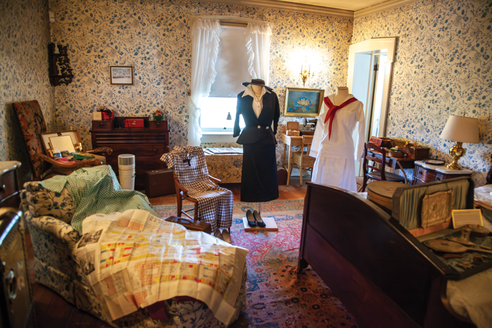 A room filled with antique furniture which includes a recliner chair with a blanket and a large folded map on top, a bed with a trunk filled with various knick knacks, a wooden chair with grey and white checkered article of clothing draped on top of it, an upholstered chair with a trunk containing picture frames in it, a chest, a dresser t with lock boxes on top, a nightstand with a lamp atop it, a writing desk with an accompanying chair, a sewing table with an accompanying chair, a red and blue rug on the floor, a wooden shelf attached to the corner of the wall, and two pictures on the wall. The smaller pictures shows a wintry scene at a house and the larger picture shows baby chickens in a hat. The walls showcase a blue and grey floral patterned wall paper with a large window being center of the photo. On the opposite wall, there is a door frame which leads into another room. 