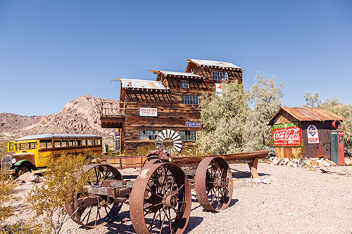 A commercial ghost town shows a school bus, remnants of a wagon, and two buildings. One building is a large barn-like wooden structure and the other appears to be a smaller outbuilding, with a Coca-Cola sign on it, and an old gas pump next to it.