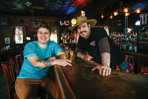 Two people smile and look at the camera, inside the Pioneer Saloon. They are at the bar - one is a bartender and the other a patron. Bottles of alcohol can be seen in the background and the bar is dark wood.