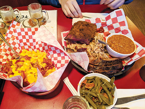 A table with bacon mac-n-cheese, brisket, pulled pork, okra, baked beans, bread, and beer.