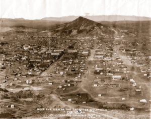 An overview shot of Goldfield in 1906. A larger downtown district can be found on center left of the image, while smaller buildings sprawl out from that area. One main large street runs through the downtown area with smaller streets criss crossing across the image. Beyond the town, a large mountain hovers over the town, while even more mountains can be seen in the distance.