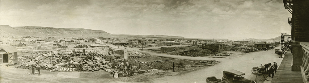 Panorama of Goldfield in 1923 after a large fire wiped out the town. Rubble where buildings once stood sprawl across the town. Roads and telephone poles can be seen amongst the rubble. On the bottom right hand corner of the image, cars and people can be seen standing next to a building. 