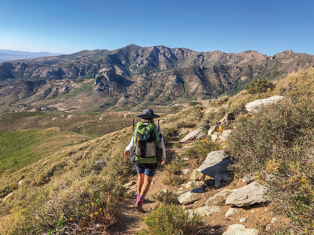 A young woman with a hiking backpack, walking on a trail leading along a mountain ridge.