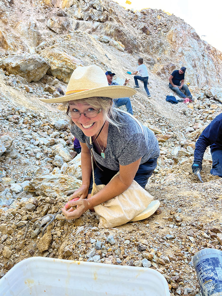 A woman digs for turquoise in the rocks and smiles at the camera.