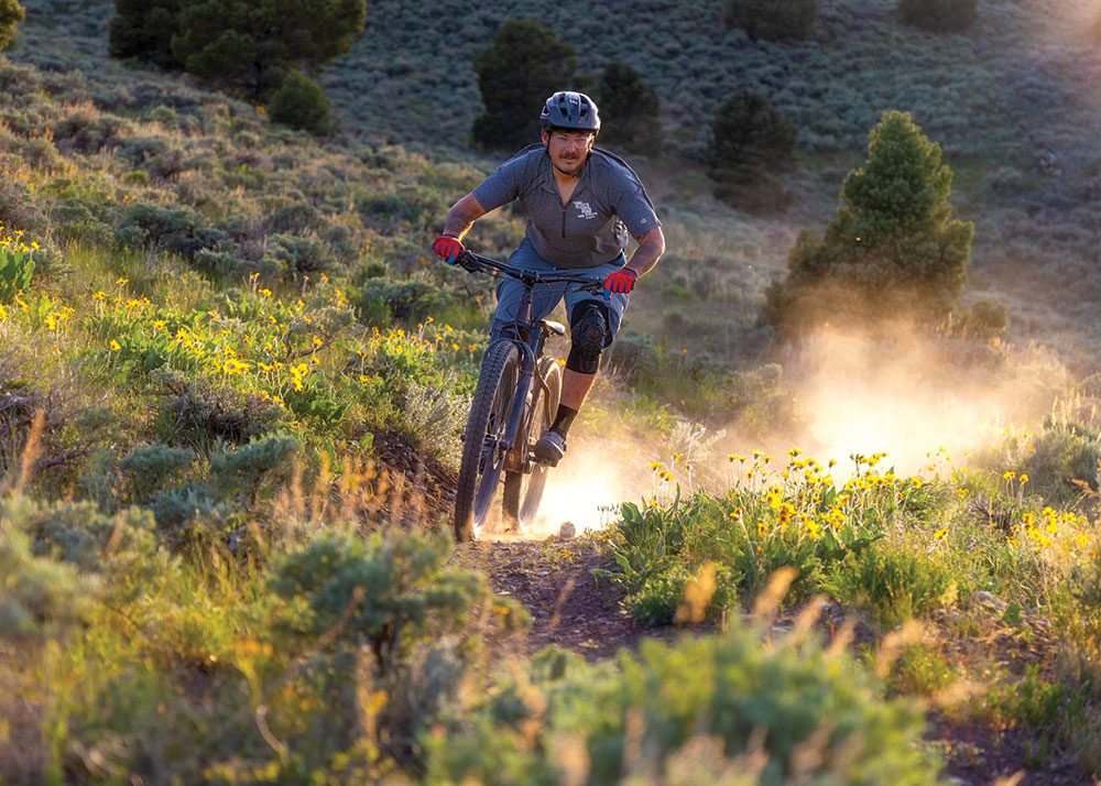 Kyle Horvath rides his bicycle through the dusty high desert, with sagebrush and flowers on either side of the trail.