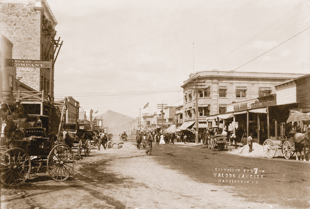 Old historic picture of Goldfield circa 1907. Buildings line a dirt road street as people are walking down and across the street. There are horse drawn carriages parked along the side of the street. Telephone poles and lines tower over the street. In the distance sits a small mountain. 