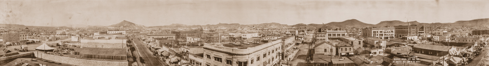A large panorama of an overview of Goldfield in 1909. Two streets split across the picture going opposite directions with buildings of various shapes and sizes lined up all along them. In the distance, mountains encase the town.